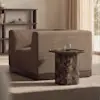 A modern living room with a brown boxy armchair, a round dark marble side table, and a glass of water. Shelves and a window are in the background, with soft natural light filling the space.