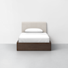 A twin bed with a beige upholstered headboard, white bedding, and a single white pillow, set on a dark wooden frame, is placed against a plain white wall on a gray floor.