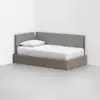 A modern daybed with a wooden base and gray upholstered backrests, placed in the corner of a minimalist room with white walls and a light gray floor. The bed has a white mattress, sheet, and pillow.