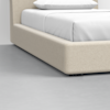 Close-up of a modern beige upholstered bed frame with white bedding and pillow, set on a smooth gray floor in a minimalist bedroom.