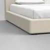 Close-up of a modern beige upholstered bed frame with white bedding and pillow, set on a smooth gray floor in a minimalist bedroom.