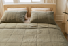 A neatly made bed with a beige headboard, two beige pillows, and a greenish-gray quilted comforter, set in a softly lit bedroom with a window and cream-colored curtains in the background.