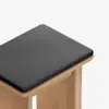 A close-up of a wooden stool with a square, dark gray cushioned seat, set against a plain white background.
