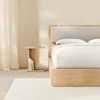 A small, modern wooden bedside table with a simple, geometric design sits next to a light wood bed frame and white bedding in a minimalistic room.