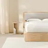 A small, modern wooden bedside table with a simple, geometric design sits next to a light wood bed frame and white bedding in a minimalistic room.
