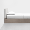 A minimalist bed with a light wood frame, a padded light gray headboard, a white mattress, white sheets, and a white pillow, set against a plain white wall on a light gray floor.