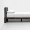 A minimalist bed with a dark upholstered headboard, white bedding, and a dark wooden frame, positioned against a white wall on a light floor.