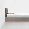 A minimalist bed with a light wood frame, a dark upholstered headboard, white bedsheets, and a pillow, set against a plain white wall and gray floor.
