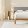 A minimalist bedroom with a wooden bed frame, light gray bedding, a matching wooden nightstand holding glassware and books, cream-colored walls, and light curtains. A soft, light rug covers part of the floor.
