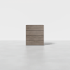 A minimalist wooden dresser with four horizontal drawers stands alone on a smooth, light gray floor against a plain white background.