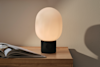 A modern table lamp with a frosted, oval-shaped glass shade and black cylindrical base sits on a wooden nightstand next to an open magazine. The lamp emits a warm, soft light.