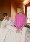 A man and a woman sit on a bed in a warmly lit room, smiling at each other. The woman wears a bright pink sweater, and the man is in a light-colored shirt. Sunlight streams in from a window behind them.