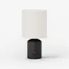 A modern table lamp with a cylindrical white fabric shade and a matte black rounded base, featuring a small circular knob on the front, set against a plain white background.