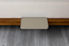 A small, rectangular beige tray is attached to the side of a bed with a wooden frame, resting above a gray wood floor. The bed has a white textured mattress.
