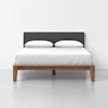A modern bed with a wooden frame, a dark gray upholstered headboard, white bedding, and two white pillows, set against a plain white wall and light-colored floor.