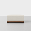 A modern, rectangular ottoman with a cream-colored upholstered top and a wooden base, photographed against a plain, light background.