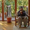 A person wearing glasses, a cap, and mismatched socks sits in a wooden chair indoors, holding a cat. Large windows and a potted plant reveal a sunlit deck and green trees outside.