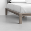 A close-up view of a modern wooden bed frame with light-colored wood, white bedding, and a gray headboard, set on a smooth, light-colored floor.