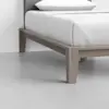 A close-up view of a modern wooden bed frame with light-colored wood, white bedding, and a gray headboard, set on a smooth, light-colored floor.