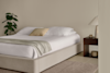 A minimalist bedroom with a beige upholstered bed, white bedding, a light throw, and a single pillow. A wooden nightstand with a lamp and decor sits beside the bed, and a potted plant is in the corner.