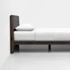 Minimalist wooden bed frame with a dark padded headboard, white mattress, white pillow, and white bedding, set against a plain white wall and floor.