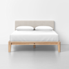 A modern bed with a light wood frame, a beige upholstered headboard, white sheets, and two white pillows, set against a plain white wall and a light gray floor.