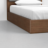 A close-up of a modern wooden bed frame with a white mattress, white bedding, and pillow, set on a light gray floor against a white wall.