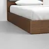 A close-up of a modern wooden bed frame with a white mattress, white bedding, and pillow, set on a light gray floor against a white wall.