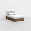 A twin bed with a wooden frame, white mattress, white pillow, and light gray upholstered headboard in a minimal, empty room with white walls and a light floor.