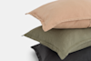 Three stacked pillows with linen pillowcases in muted colors: peach on top, green in the middle, and dark gray at the bottom, against a plain white background.
