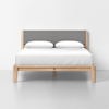A modern wooden bed frame with a gray upholstered headboard, white bedding, and two white pillows, set against a plain white wall in a minimalist room.