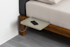 A small, light gray bedside shelf attached to a wooden bed frame holds a smartphone connected to a charging cable. The bed has a beige mattress and bedding, and the floor is light gray wood.