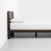 A side view of a neatly made bed with a dark wooden frame and headboard, white bedding, and a pillow, positioned against a white wall in a minimal, modern room.