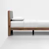 A minimalist wooden bed frame with a gray upholstered headboard, white bedding, and a single pillow, positioned against a white wall in a bright, uncluttered room.