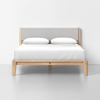 A modern wooden bed frame with a light gray upholstered headboard, white pillows, and a neatly made white bedspread, set against a plain white wall and floor.