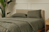 A neatly made bed with olive green bedding and two pillows stands next to a wooden nightstand and a tall green plant in a minimalist, light-colored room.