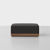 A modern, rectangular ottoman with a dark gray upholstered cushion and a wooden base, placed on a light-colored floor against a plain white background.