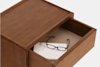 A wooden nightstand with an open drawer containing a closed book and a pair of eyeglasses with dark frames.