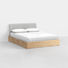 A modern bed with a light wooden frame, a gray upholstered headboard, and white bedding with two pillows, placed in a minimalistic, bright room with white walls and flooring.