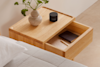 A light wooden nightstand with an open drawer revealing two notebooks and a pen. On top, there is a plant in a ceramic vase and a black cylindrical container labeled THUMA. A bed with a grey quilt is next to the nightstand.