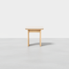 A small, light wood square stool with simple, clean lines, positioned on a smooth, light gray floor against a plain white background.