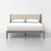 A modern bed with a light wood frame, beige upholstered headboard, white bedding, and two white pillows, set against a plain white wall and floor.