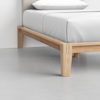Corner view of a modern wooden bed frame with light wood legs and a white mattress, set on a smooth, light gray floor against a plain white wall.