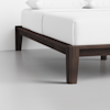 A close-up of a modern bed with a dark wooden frame and white bedding, showing the corner and leg of the bed against a light-colored floor and wall.