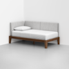 A minimalist daybed with light gray upholstered back and side panels, a white mattress, a single white pillow, and a wooden frame, placed in the corner of a bare room with light gray flooring and white walls.