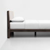 A modern bed with a dark wooden frame, white mattress, pillow, and bedding, set against a plain white wall and floor. The design is minimalistic and clean.