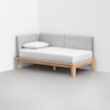 A minimalist daybed with light gray upholstered backrest and sides, a white mattress and pillow, and a natural wood frame, placed in the corner of a plain, light-colored room.