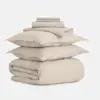 A neatly stacked set of beige bedding, including a folded duvet, two pillows with pillowcases, a flat sheet, and two folded pillow shams, all arranged on a white background.