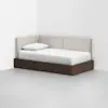 A modern daybed with a dark wooden frame and beige upholstered backrest is placed in the corner of a minimalist, white-walled room. The bed is made with white bedding and a single pillow.
