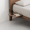 A wooden bedside platform attached to a bed holds a black cup and saucer, a notebook, and a pen. The bed has beige bedding and the floor and wall are light-colored. The scene is minimalistic and modern.